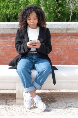 Bukleli genç siyahi bir kadın bankta oturuyor dışarıda cep telefonu kullanıyor tuğlalara ve ağaçlara karşı. Dikey fotoğraf.