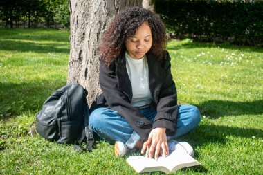 Kıvırcık saçlı siyah bir kız, yanında sırt çantasıyla çimenlere yaslanan bir kitap okuyor. yatay resim.