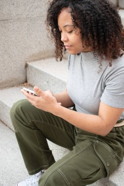 Kıvırcık saçlı bir kadın merdivenlerde otururken cep telefonu kullanıyor..