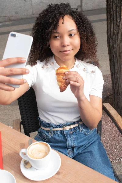 Excited black woman with croissant and smartphone taking a selfie while having breakfast in a coffee shop. Lovely African American lady taking a photo, creating happy memories in a restaurant.