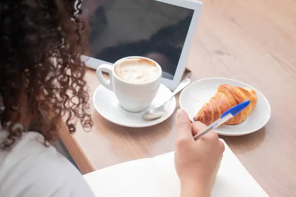 Kahve içerken ve kruvasan molası verirken tabletini kullanarak masanın üzerine not defteri yazan bir kız..