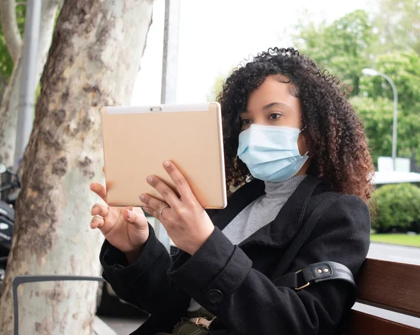 Siyah, kıvırcık saçlı, maske takan ve tablet giyen bir kız..