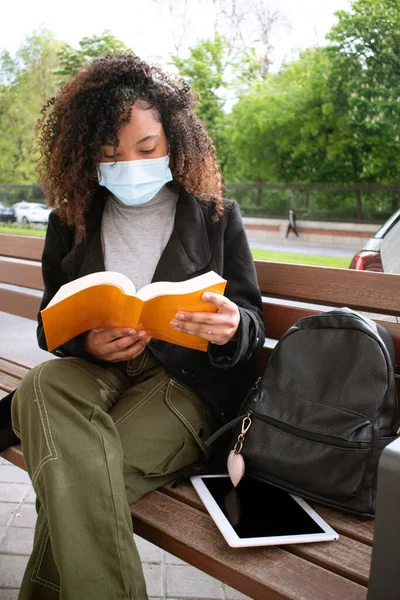 Kıvırcık saçlı, yüz maskesi takan ve yanındaki bankta turuncu kitap okuyan genç bir kadın sırt çantası ve bir tablet..