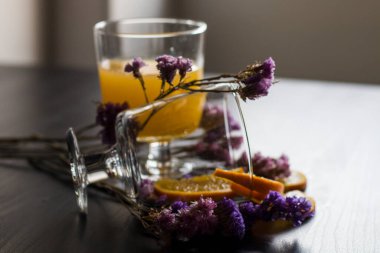 Bir bardak hamurlu ve dilimlenmiş meyveli sağlıklı portakal suyu ahşap zemin üzerinde izole edilmiş güneş ışığı ve mor çiçekli süslemeler. Portakal suyu ve taze meyveler masada..