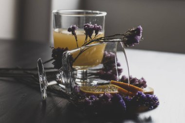 Bir bardak hamurlu ve dilimlenmiş meyveli sağlıklı portakal suyu ahşap zemin üzerinde izole edilmiş güneş ışığı ve mor çiçekli süslemeler. Portakal suyu ve taze meyveler masada..