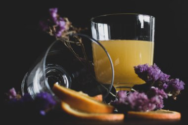 Bir bardak sağlıklı portakal suyu hamurlu ve dilimlenmiş meyve mor çiçeklerle siyah arka planda izole edilir. Portakal suyu ve taze meyveler masada..