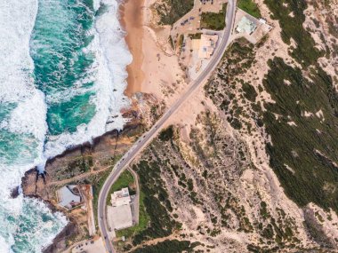 Atlantik Okyanusu 'na bakan vahşi güney kıyı şeridi boyunca giden araçların bulunduğu dar bir yolun hava manzarası kayalıklarda, Cascais, Portekiz.