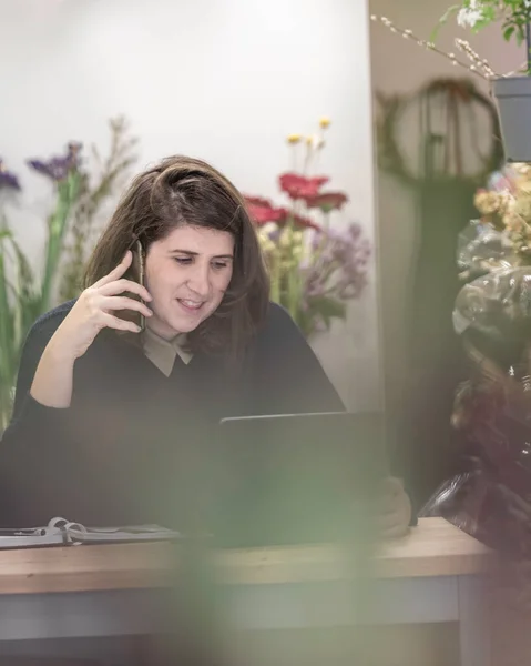 View of a young woman working as florist in a flower shop and talking on the phone with clients. Young entrepreneur concept.