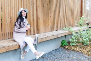 Elinde bir fincan kahve olan güzel bir kadın çitin yanında oturuyor.