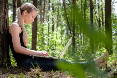 girl in the forest sits leaning against a tree, working on a laptop. rest at nature. in the background blurred silhouettes of trees. digital detox. Mental health