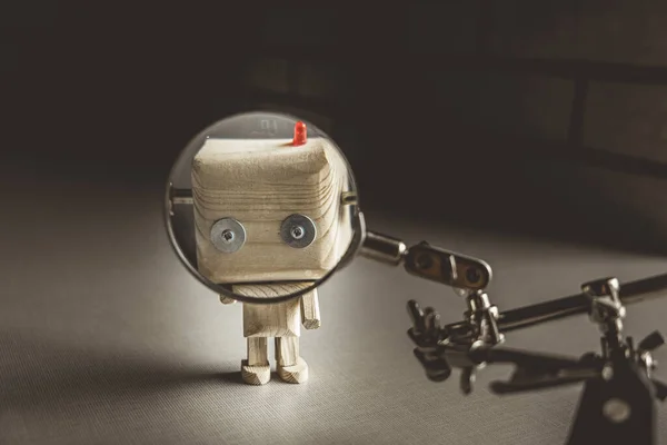 wooden robot android stands on a table under a magnifying glass. metal clamps with a magnifying glass. the face of a robot in the beautifying glass. human study concept