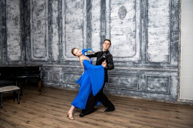 Genç adam, gri dans salonunda mavi balo elbiseli bir kızla dans ediyor.