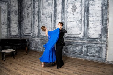 Genç adam, gri dans salonunda mavi balo elbiseli bir kızla dans ediyor.