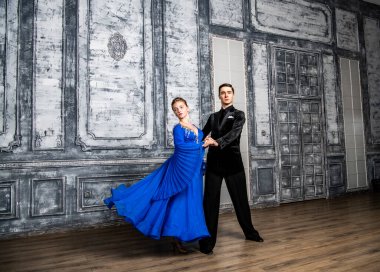 Genç adam, gri dans salonunda mavi balo elbiseli bir kızla dans ediyor.