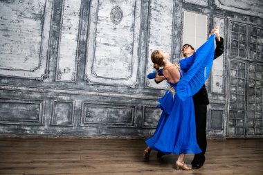 Genç adam, gri dans salonunda mavi balo elbiseli bir kızla dans ediyor.