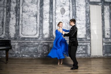 Genç adam, gri dans salonunda mavi balo elbiseli bir kızla dans ediyor.