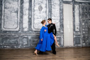 Genç adam, gri dans salonunda mavi balo elbiseli bir kızla dans ediyor.