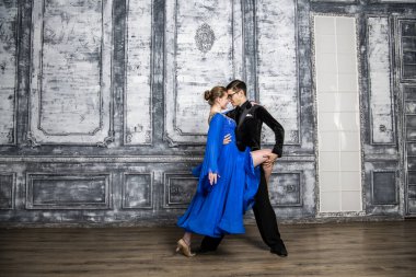 Genç adam, gri dans salonunda mavi balo elbiseli bir kızla dans ediyor.