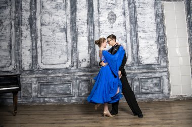 Genç adam, gri dans salonunda mavi balo elbiseli bir kızla dans ediyor.