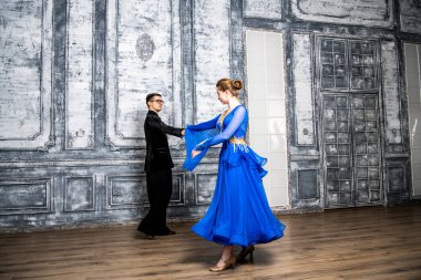 Genç adam, gri dans salonunda mavi balo elbiseli bir kızla dans ediyor.