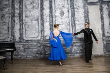 Genç adam, gri dans salonunda mavi balo elbiseli bir kızla dans ediyor.