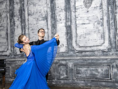 Genç adam, gri dans salonunda mavi balo elbiseli bir kızla dans ediyor.