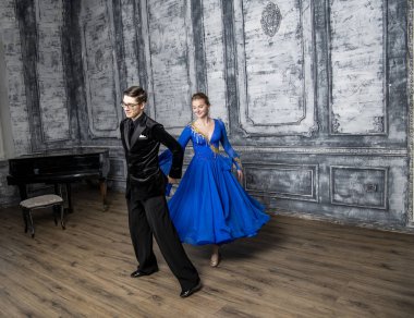 Genç adam, gri dans salonunda mavi balo elbiseli bir kızla dans ediyor.