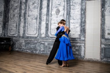 Genç adam, gri dans salonunda mavi balo elbiseli bir kızla dans ediyor.