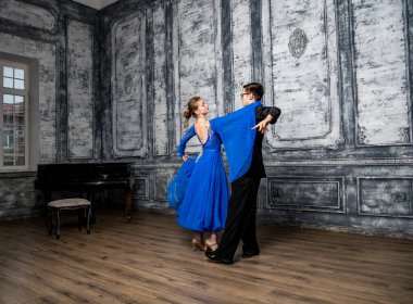 Genç adam, gri dans salonunda mavi balo elbiseli bir kızla dans ediyor.