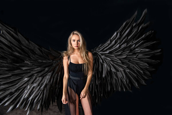Beautiful blonde girl in a black suit with black wings on a black background