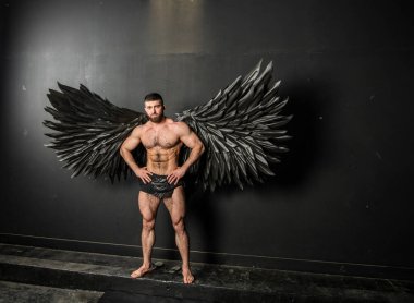Siyah arka planda siyah kanatları olan melek kostümlü yakışıklı kaslı adam.