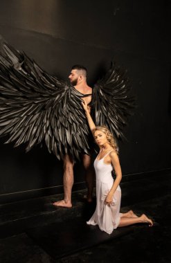 handsome muscular man in angel costume with black wings and fragile defenseless beautiful girl in white clipart