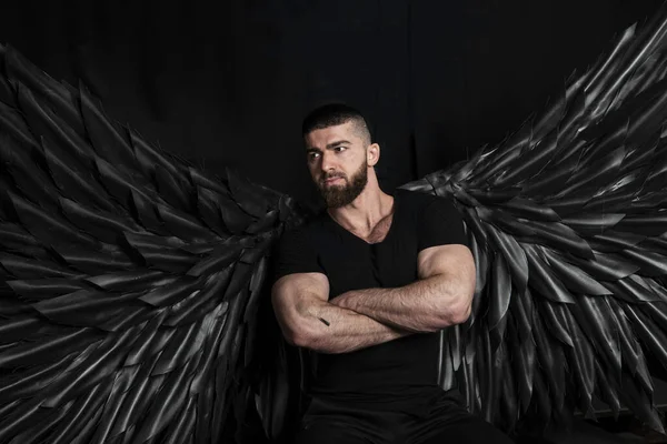 Siyah arka planda siyah kanatları olan melek kostümlü yakışıklı kaslı adam.