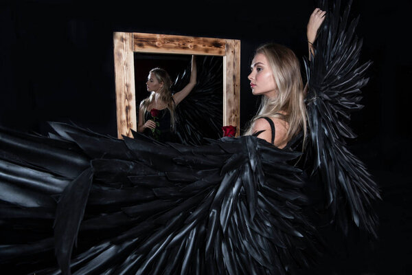 beautiful girl dressed as an angel with black wings near the mirror on a black background
