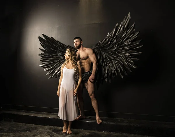 handsome muscular man in angel costume with black wings and fragile defenseless beautiful girl in white