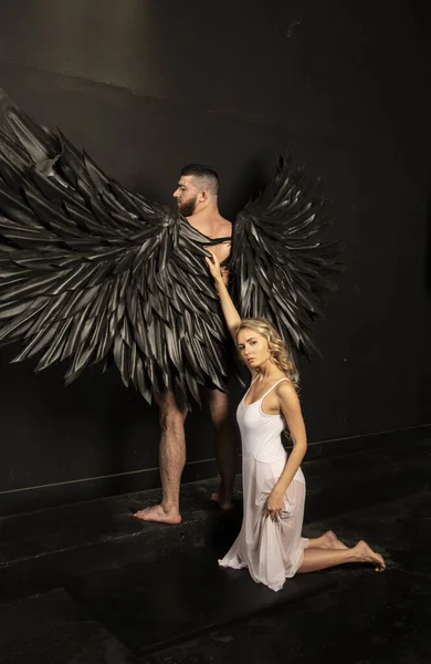 handsome muscular man in angel costume with black wings and fragile defenseless beautiful girl in white