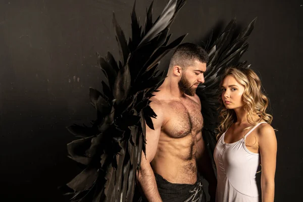 handsome muscular man in angel costume with black wings and fragile defenseless beautiful girl in white