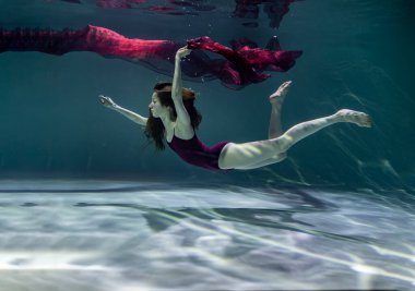   Mavi arka planda kırmızı bir pareo ile su altında poz veren bordo mayo giymiş güzel bir kız.