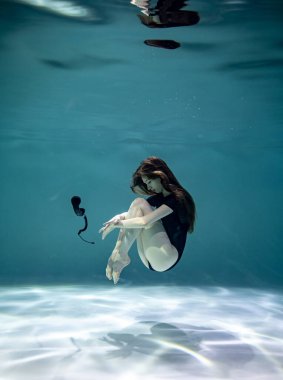 Siyah mayo giymiş, mavi arka planda siyah bir telefon ahizesiyle su altında poz veren güzel bir kız. 