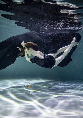Siyah mayo giymiş güzel bir kız ve siyah pareo su altında mavi bir arka planda akvaryumda balıklarla oynuyor.
