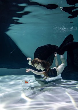 Siyah mayo giymiş güzel bir kız ve siyah pareo su altında mavi bir arka planda akvaryumda balıklarla oynuyor.