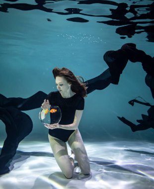 Siyah mayo giymiş güzel bir kız ve siyah pareo su altında mavi bir arka planda akvaryumda balıklarla oynuyor.