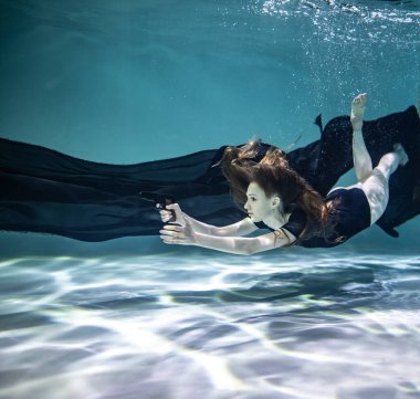 Siyah pareolu siyah mayo giymiş güzel bir kız mavi arka planda, su altında elinde siyah bir tabanca tutuyor.