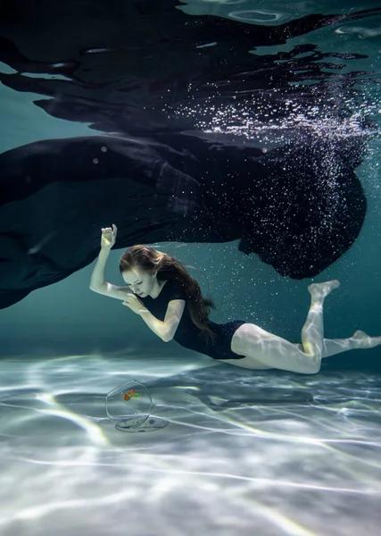 Siyah mayo giymiş güzel bir kız ve siyah pareo su altında mavi bir arka planda akvaryumda balıklarla oynuyor.
