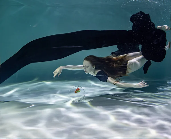 黒のパレオと黒の水着の美しい女の子が水中の青の背景に水族館で魚と遊ぶ — ストック写真