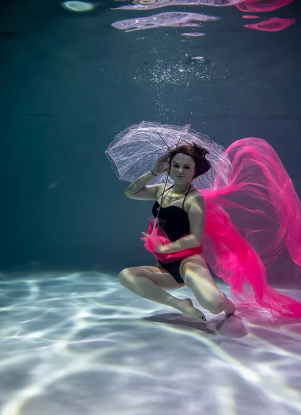 青い背景に赤いパレオとピンクの傘を持った黒い水着の美しい女の子が水中で — ストック写真