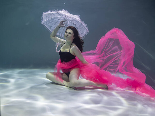 beautiful girl in a black swimsuit with a red pareo and a pink umbrella on a blue background underwater