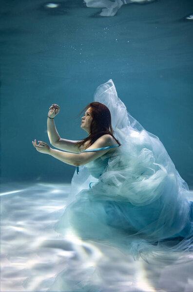 beautiful girl with red hair in a ball gown blue dress underwater on a blue background