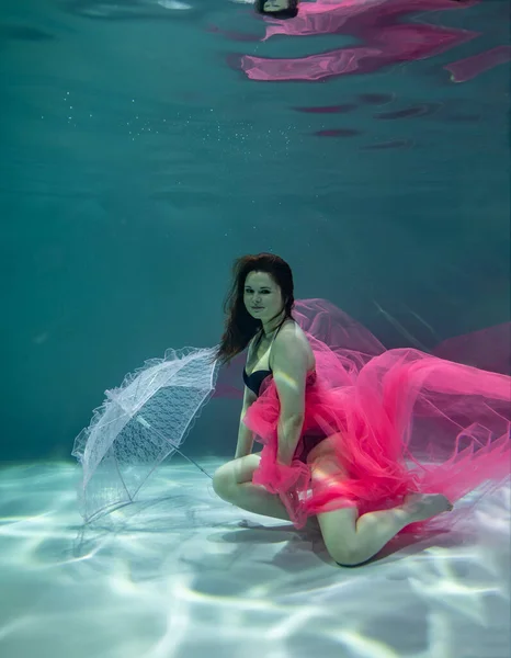 黒の水着とピンクのパレオと青い背景にピンクの傘を水中に持つ美しい女の子 — ストック写真