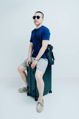 Koyu mavi tişörtlü genç bir adam siyah bir bavulun üzerinde oturuyor.